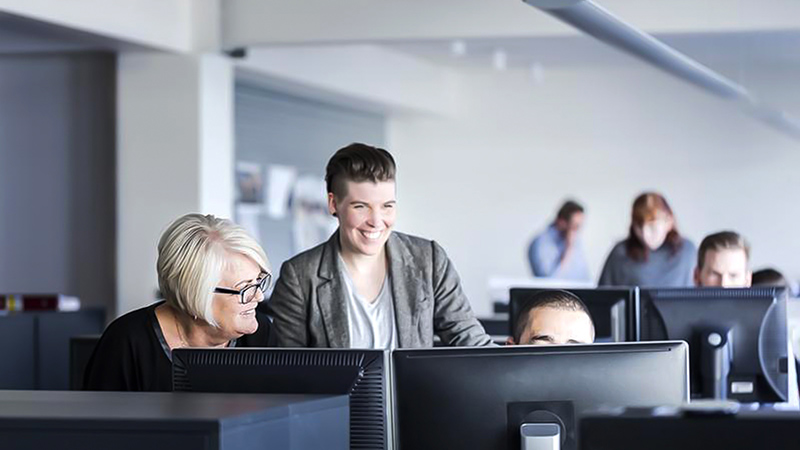 Happy coworkers gather around a teammate's desk and discuss content on his computer. 