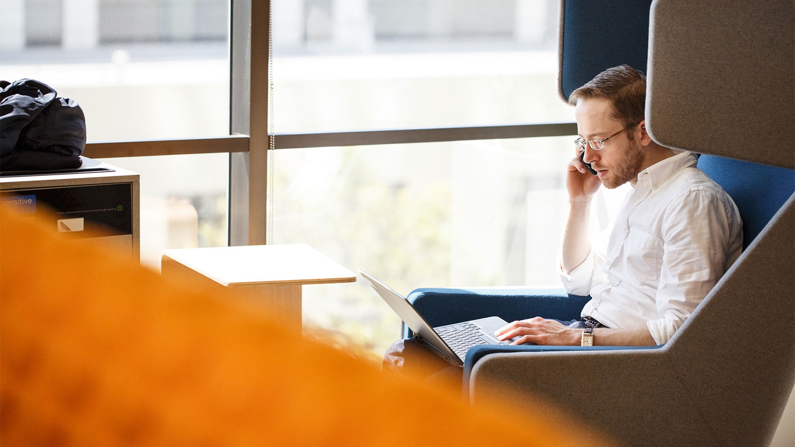 Man sitting on a chair in office environment, talking on his cell phone while looking at his laptop. Man sitting on a chair in office environment, talking on his cell phone while looking at his laptop.